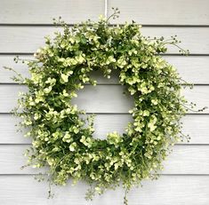 a green wreath hanging on the side of a white building with flowers in it and greenery