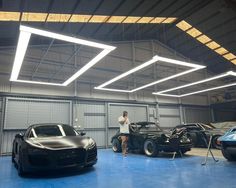 a man standing next to two cars in a garage with overhead lighting above the cars