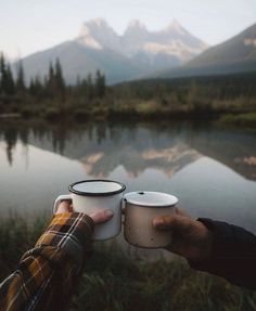 two people holding coffee cups in their hands near a lake with mountains in the background