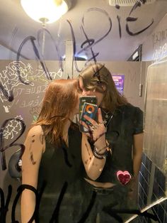 two women taking a selfie in a bathroom mirror with graffiti all over the walls