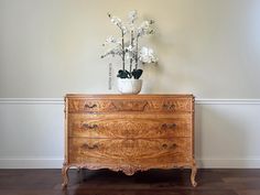 a vase with flowers on top of a wooden dresser next to a white wall and hardwood floor