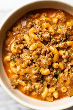 a white bowl filled with pasta and meat in tomato sauce on top of a marble table