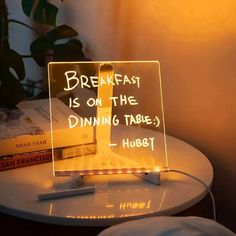 a light up sign that reads breakfast is on the dining table hubet next to some books