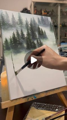 a person is holding a paintbrush and painting a picture on an easel with trees in the background