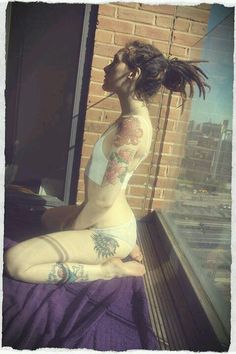 a woman sitting on top of a bed in front of a window with tattoos on her arms