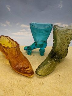 three glass vases sitting next to each other on a sandy surface with clouds in the background