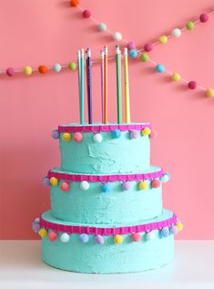 a three tiered blue cake with multi colored candles on top and pom - poms around the edges