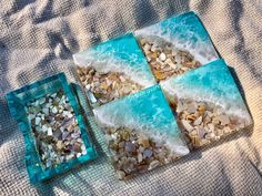 three square glass coasters with sea shells and sand in them sitting on a bed