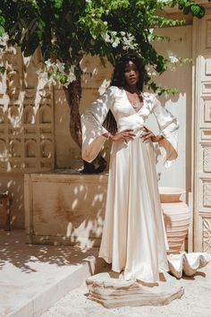 a woman standing in front of a tree wearing a white dress with long sleeves and bell sleeves