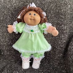 a cabbage doll with brown hair and green dress sitting on the ground next to a gray carpet