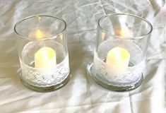 two clear glass candles are sitting on a white sheet with lacy doily around them