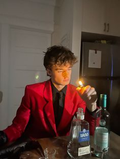 a man in a red suit holding a lit candle at a table with bottles and glasses on it