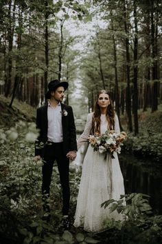 a bride and groom holding hands walking through the woods with flowers in their hair, dressed in black