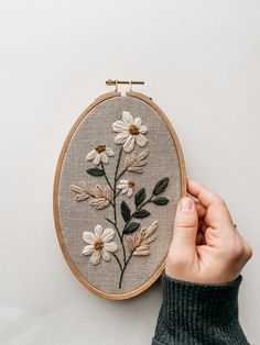 a person holding up a hand embroidered flower on a wall mounted embroidery hoop with white daisies and green leaves