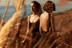 two women standing next to each other in tall grass near the ocean, one with her back turned towards the camera