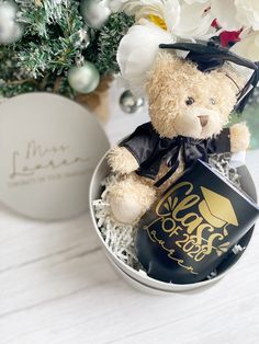 a teddy bear in a graduation hat and gown sits inside a small gift box next to a christmas tree