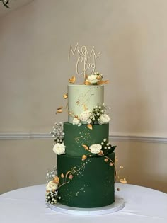 a green wedding cake with white flowers and gold lettering on top is sitting on a table