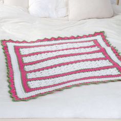 a crocheted pink and white blanket on a bed with pillows in the background