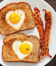 two pieces of toast with eggs in the shape of heart and bacon on top, next to orange juice