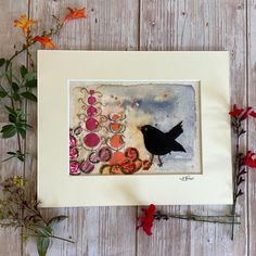 a black bird sitting on top of a wooden table next to flowers and plants in a white frame