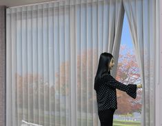 a woman standing in front of a window looking out the window with white vertical blinds