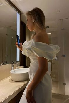 a woman in a white dress looking at her cell phone while standing in front of a bathroom mirror