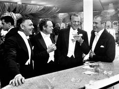 four men in tuxedos standing at a bar laughing and drinking wine while another man looks on