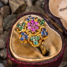 This vintage floral ring features clusters of emeralds, rubies, and sapphires that are accented with a gold rope motif. The ring is crafted in 18k yellow gold with a rich patina. It is currently a size 6.25. Several of the stone have a bit of light wear. Antique Multi-stone Yellow Gold Cluster Ring, Antique Multi-stone Cluster Ring In Yellow Gold, Yellow Gold Multi-stone Cluster Sapphire Ring, Yellow Gold Cluster Multi-stone Sapphire Ring, Formal Multi-stone Yellow Gold Flower Ring, Vintage Yellow Gold Flower Ring With Gemstone, Cluster Emerald Ring In Yellow Gold With 17 Jewels, Vintage Style Emerald Cluster Ring In Yellow Gold, Heirloom Yellow Gold Cluster Ring With Multi-stone