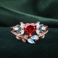 a red heart shaped ring with leaves and stones on the side, sitting on a green velvet surface