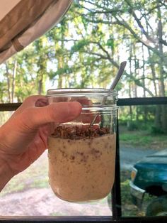 a person holding a spoon in a glass jar with food inside it and trees outside the window