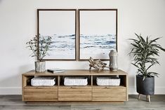 two framed pictures hang on the wall next to a wooden cabinet with baskets and plants