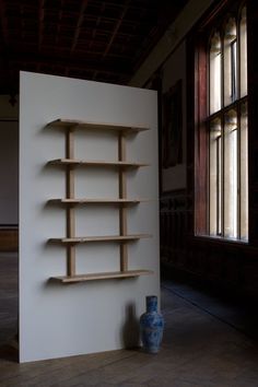 a wooden shelf sitting next to a vase on the floor in front of a wall