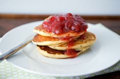 stack of pancakes covered in strawberry jam on a white plate with a fork and spoon