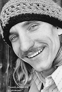 a man with long hair wearing a knitted hat smiles at the camera while standing in front of a wooden fence