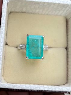 an emerald and diamond ring sitting in a white box on top of a cushioned surface