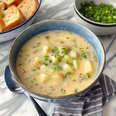 a bowl of potato soup next to two bowls of bread and green onions on a marble table