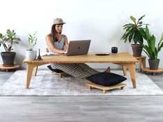 a woman sitting at a wooden table with a laptop on her lap and potted plants in the background