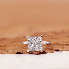 a princess cut diamond ring sitting on top of a piece of wood with leaves around it