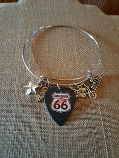 "Fun charms and a Route 66 guitar pick make up this stainless adjustable bangle bracelet. The bangle expands to slide over the hand but fits a 7\" wrist best" Adjustable Metal Bracelets For Concerts, Adjustable Bracelet Jewelry For Concerts, Adjustable Music-themed Jewelry For Concerts, Adjustable Silver Bracelets For Concerts, Adjustable Silver Jewelry For Concert, Adjustable Silver Jewelry For Concerts, Adjustable Metal Music-themed Jewelry, Silver Bracelet For Concerts, Silver Bracelet Jewelry For Concert