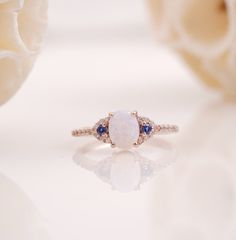 an opal and diamond ring sits on a white surface next to some flower petals