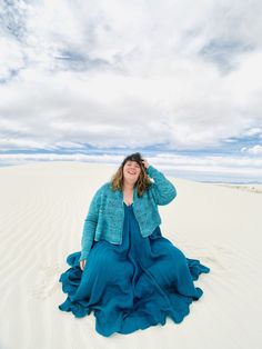 a woman sitting on top of a sandy beach under a blue sky with white clouds