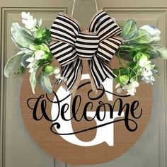 a welcome sign hanging on the front door with flowers and greenery attached to it