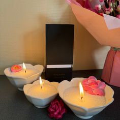 three white bowls filled with candles next to a pink rose and a black box on a table