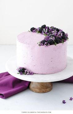 a pink cake with blackberries on top and purple flowers in the middle is sitting on a white plate