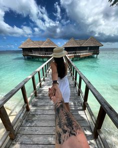 a man and woman holding hands while standing on a wooden bridge over water with thatched huts in the background