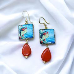 "The Sicilian earrings with prickly pear decoration on ceramic and lava stone tile are a beautiful example of traditional Sicilian art. Lava stone is a volcanic stone typical of the Sicilian region and was used to create a square base for setting prickly pears in white ceramic. The soutache technique, which is used to create the golden frame around the tile, consists in setting a metal cord in order to create a weave that surrounds the stone. This diamond effect gives the earrings an elegant and refined look. The decoration of prickly pears is a symbol of Sicily and its culture, as this plant is widespread in the region. The earrings with prickly pears on ceramic and lava stone therefore represent a tribute to the beauty and tradition of Sicily. You can choose between the two variants prop Sicilian Art, Tile Earrings, Volcanic Stone, Golden Frame, Stone Tile, Prickly Pear, Lava Stone, Gold Plated Silver, Stone Earrings