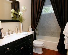 a white toilet sitting next to a bath tub in a bathroom under a window with black curtains