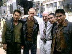 four men are posing for the camera in front of a building and one man is wearing a leather jacket