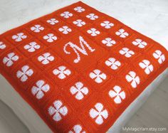 an orange crocheted blanket with white flowers and the letter m on it is sitting on a bed