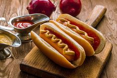 two hotdogs with ketchup and mustard on a cutting board next to apples